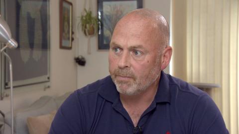 Jim Newman, wearing a dark blue polo shirt, sits in his living room. He has a straight face as he looks off camera. There is a light grey sofa and artwork on the walls in the background