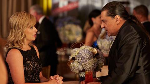 An image of Amy Poehler and Jonathan Joss sitting at a table. Amy is wearing a lace black dress and Jonathan is wearing a shiny black suit with his long brown hair tied back in a ponytail. 