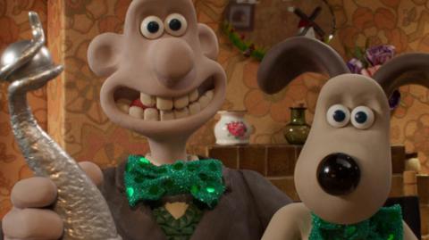 Image of clay modelled characters Wallace and his dog Gromit. Wallace is holding a silver trophy. They are both wearing green shiny green bow ties. In the background can be seen a tile fireplace, oval mirror above the fire and a china and glass vase.