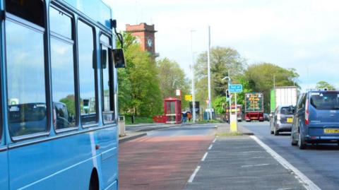 A bus, using a bus lane on the A64, approaches a red bus shelter where a person is waiting to board.