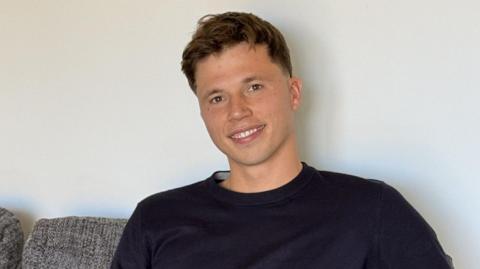 Freddie Woodward: a young man with short dark hair sits on a grey sofa, smiling at the camera. He has short brown hair and wears a navy jumper.