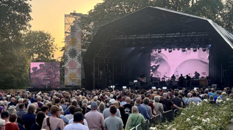 A concert taking place in York's Museum Gardens at dusk. A crowd is gathered in front of a stage surrounded by trees. The audience fills most of the foreground, with hundreds of people standing shoulder-to-shoulder facing the performance.