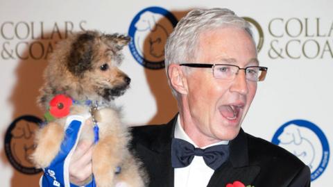 A silver haired Paul O'Grady wearing glasses and a black jacket with bow tie, carrying a dog with a blue collar and both are wearing red poppies. They are standing infront of a sign at a press-call 