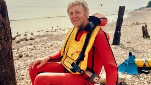 A man with fair hair sitting on a pebble beach. He has a red wetsuit on and a yellow life vest over the top. He is smiling as he looks into the camera, sideways facing.