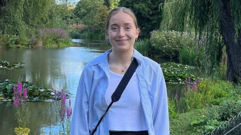 Libby smiling into the camera in front of a pond with lily pads, plants and flowers. She has blonde hair which is tied back and is wearing a blue and white striped shirt with a white tank top underneath. She is also wearing a black cross-body bag and a simple silver necklace which is silver with a small pendant.