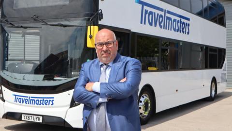 A man in a blue suit wearing glasses with his arms crossed, looking stern. He is stood in front of a white bus with a blue logo reading "Travelmasters" on the front and the side.