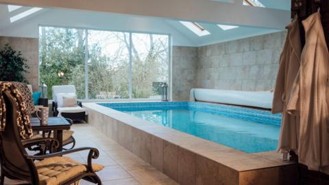 A generic image of an indoor home swimming pool in a bright room with poolside loungers, armchairs and large windows.