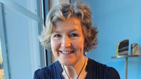 A woman in a navy blue dress smiling at the camera while sitting in front of a blue wall. 