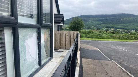 A side-on view of the exterior of the house in Mullaghbawn where a pipe bomb exploded overnight. Two of the front windows are shattered and a piece of wood or cardboard has been used to board up the broken window.  There is an empty road in front of the house and a mountain in the background. 