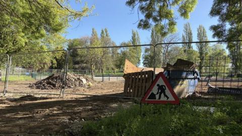 A metal fence surrounds a construction area in a park. A mound of earth is visible alongside a large skip filled with waste. Leafy trees, grass, and a paved path surround the area. There is also a clear blue sky.