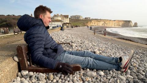 A man is sat on a bench submerged in stones on a beach. He is wearing a blue coat and gloves with white shoes. There are cliffs in the background and the sky is blue.