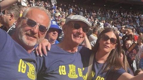 Julian Wantling with a man and a woman inside Wembley in 2025. They are wearing blue Southend United T-shirts and sunglasses.