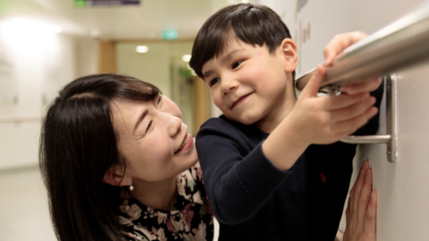 Mariko and Joshua inside a corridor at Great Ormond Street Hospital. They are looking at each other and smiling.