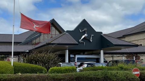 The exterior of Noble's Hospital, the pointed roof of the foyer has a silver metal three legs of man sculpture. Cars are parked outside, a Manx flag blows from a flag pole.