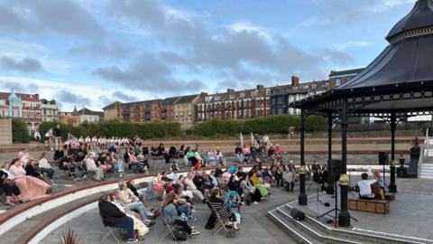 Dozens of people sat in chairs facing a black bandstand. There are green hedges and taller buildings in the background. 