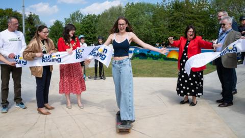 A young woman wearing jeans and a blue top skateboarding through a ribbon which has "Luton Rising" on it and is being held by men and women. She is in a skatepark with trees in the background.