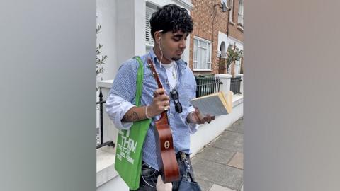 A young man poses on a street as he reads a feminist book in one hand and clutches a ukulele in the other. He has a tote bag on his shoulder as well as a Labubu and vintage cap hanging from his belt loops.