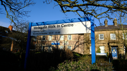 A blue and white sign reading: "Westgate Walk In Centre" sits in front of two, two-storey buildings.