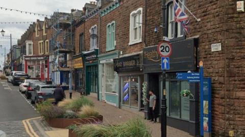 A view of the shops on Senhouse Street in Maryport. A range of shops can be seen and planters with different types of grass growing in them can be seen on the pavements in front of the shops.