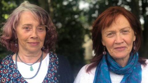 A close-up image showing the heads and shoulders of Margaret and Paula Martin. They are both older women who are looking directly into the camera with a neutral expression. On the left, Margaret has short, purple hair with grey roots and she is wearing a layered necklace and a flower patterned top. On the right is Paula, who has longer, auburn hair. She is wearing a blue pattern scarf, a white top and a pair of dangly earrings. They are stood in front of a grouping of trees but the background is blurred.