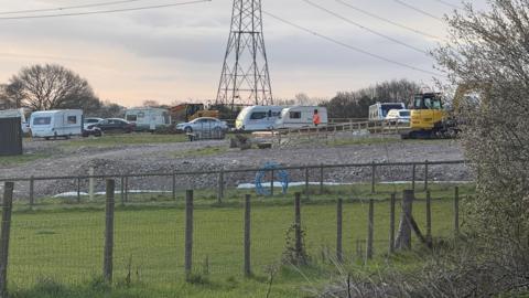 A view of the field. About six caravans are parked on hardstanding along with other vehicles and yellow diggers. There is a pylon in the middle of the field. 
