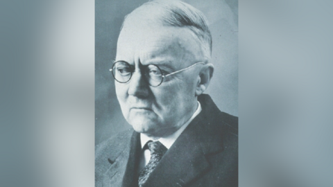 A black and white photograph of a man in a suit with short hair wearing round wire frame glasses. He is looking off to the side.