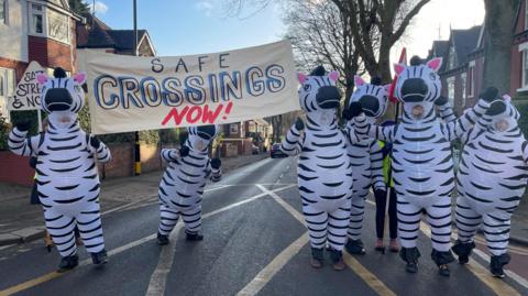 A number of people in inflated zebra costumes are stood in a box junction on the road. They're carrying a banner which reads: "Safe Crossings Now!"