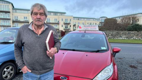 Wayne Stokes pictured with short grey hair and a moustache. He is wearing a grey quarter-zip knitted jumper which has a salmon pink collar poking out the top. He has his hand tucked into the pocket of his blue denim jeans and is clutching a burgundy notebook in his other hand. Wayne stands in front of a burgundy Ford Fiesta which has the name of his driving school 'Roadcraft' on the top with a red 'L'