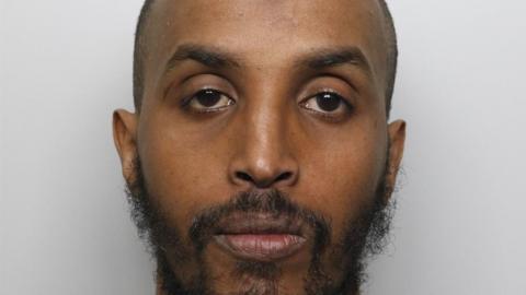 A mugshot of a bald man with a thin mid-length black beard. He has dark eyes and arched eyebrows. He has a neutral expression.