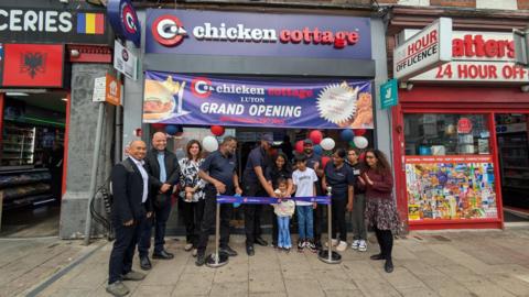A group of people, some wearing uniforms for a chicken shop, and others smartly dressed, are celebrating outside a new Chicken Cottage chain