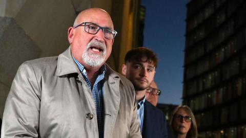 Radd Seiger is pictured standing outside a court building before a bank of microphones. He is tall, bald, with a short grey beard and is wearing dark-framed glasses and a beige raincoat over an open-necked blue and white checked shirt. He is flanked by several members of Harry Dunn's family who are looking on.
