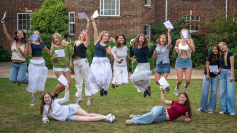 A group of young women are all holding pieces of paper with their results, smiling at the camera. Some are jumping in the air while others are lying on the ground posing.