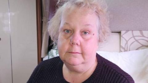 Donna is sitting in a motorhome-style space. She has light, slightly unkempt hair and wears a dark knitted jumper. In the background, there are white cushions, wood‑panelled cupboards and cream-coloured storage units.
