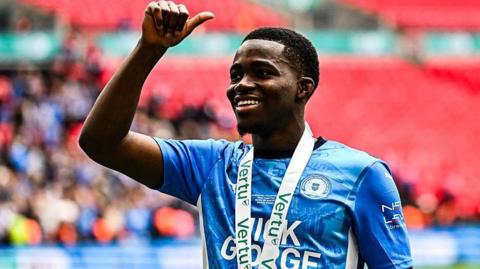Kwame Poku giving the crowd a thumbs-up. He is wearing a blue Peterborough shirt and has a medal around his neck