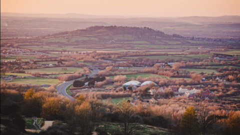 Chosen Hill from a high viewpoint. There is a dual carriageway running along to the left and in the centre a large green hill covered in trees and hedges.