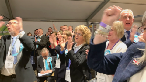 Lots of people with their hands in the air celebrating, most of them in black suits with turquoise rosettes.