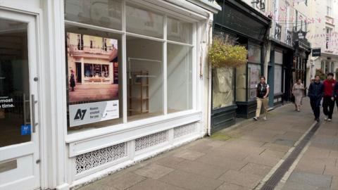 Empty shop fronts in Mill Street, St Peter Port, Guernsey. People can be seen walking on the right side of the image while a white empty shop is on the left with a boarded up green shop front next to it
