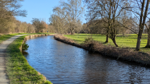 A towpath is on the far left of the photo, by grass, which is next to the canal. A field is to the right of the canal and trees are to the left and right of the canal.
