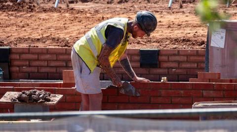 A construction worker in a yellow hi-vis vest and grey shorts is laying red bricks on a partially built brick wall, using a trowel to apply mortar.