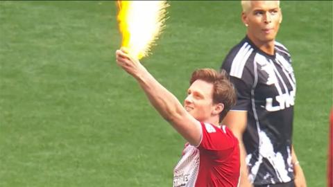 Max Fosh holds up a yellow card with sparks flying out of it on a football pitch 