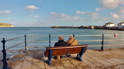 Two people are sat on a bench looking out over the sea. The couple have their back to the camera. A metal railing runs in front of the bench. It is a sunny day with a few wispy clouds in the sky.