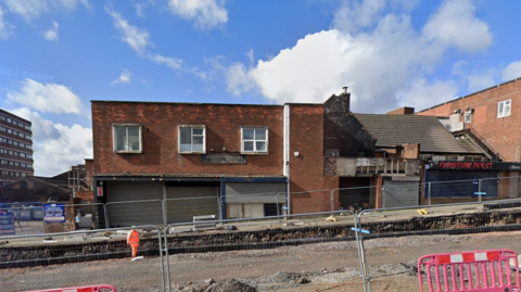 A brick two-storey building with roadworks outside the front. The road has been dug up and metal fencing is on either side. 