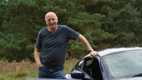 A smiling Martin Druce stands at the open door of his polished blue BMW car. One hand is on the roof of the car and the other is in his pocket. He has short white hair and is wearing a black t-shirt and blue jeans.
