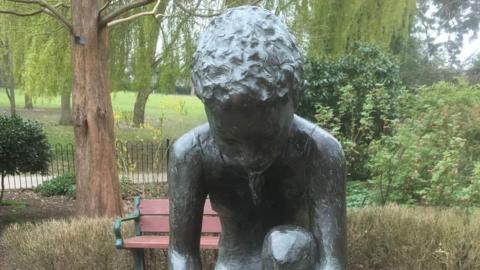 The "Child in the Park" statue in situ in Lake Meadows. It is a figure of a child crouching. A bench, a fence and greenery can be seen behind it.