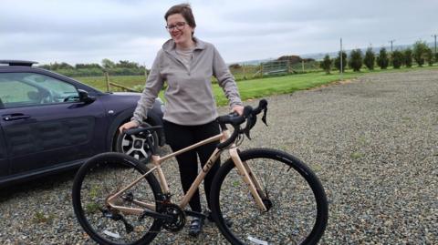 Jodie pictured with a gold coloured race bike wearing black leggings and a beige jumper. She has short dark hair and wears glasses and is standing on a gravel car park next to a black car, with fields in the background