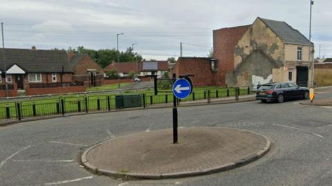 A roundabout connecting High Street and Elemore Lane in Sunderland. A black car is travelling away from the camera. A number of brick buildings can be seen to the side of the road.