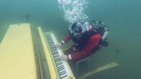 Joe Jenkins wearing diving gear whilst underwater playing a yellow piano