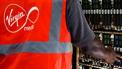 A Virgin media installer - identified by the logo on the back of his high-viz vest - reaches across to touch the electrical innards of a telecoms cabinet