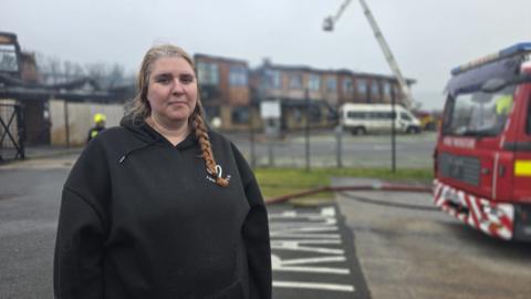 A woman called Elizabeth stands next to a red fire engine as firefighters on an aerial ladder platform in the distance continue to battle a blaze at a school in the background. Elizabeth is wearing a black hoodie and has long hair tied into a braided ponytail, which is draped over her left shoulder.