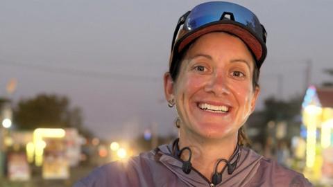 A female runner stands outside at dusk, wearing a cap, running glasses on her head and a purple waterproof. She is smiling at the camera. In the background there are dim lights.
